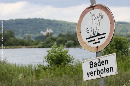 Ein Schild mit der Aufschrift "Baden verboten" und einem Symbol, das eine Person zeigt, die ertrinkt. Im Hintergrund sieht man einen See, umgeben von Bäumen und Hügeln.