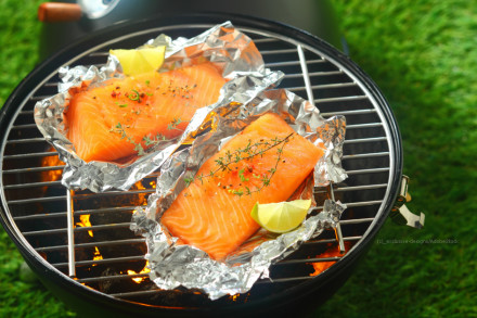 Zwei Lachsfilets liegen in Aluminiumfolie auf einem Grillrost. Sie sind mit frischen Kräutern und Gewürzen garniert. Eine Zitronenscheibe befindet sich auf jedem Lachsfilet. Der Grill steht draußen auf grünem Gras.