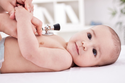Ein Baby liegt auf dem Rücken und wird von einem Erwachsenen, der ein Stethoskop benutzt, untersucht. Das Baby trägt nur eine Windel.