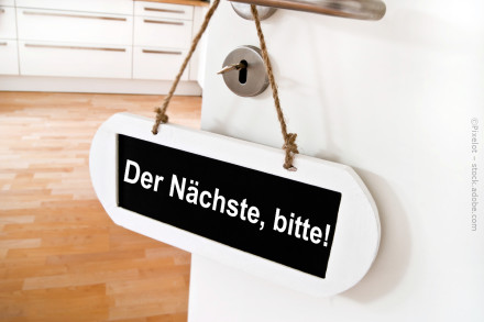 Ein Schild mit der Aufschrift "Der Nächste, bitte!" hängt an einer Tür. Im Hintergrund ist eine moderne Küche mit weißen Schränken und Holzfußboden zu sehen.