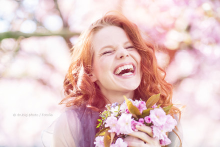 Eine Frau mit langen, lockigen, roten Haaren lacht herzlich und hält einen Strauß pinker Blumen. Der Hintergrund ist mit verschwommenen Pastellfarben gesättigt und deutet auf einen blühenden Garten oder Park hin.
