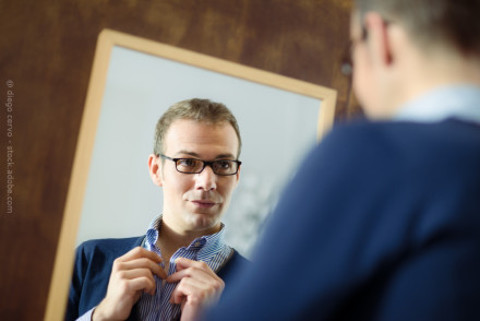 Ein Mann mit Brille steht vor einem Spiegel und knöpft sein Hemd zu. Er trägt ein blau-weiß gestreiftes Hemd und ein dunkelblaues Oberteil. Die Szene wird in einem Innenraum mit einem hölzernen Hintergrund dargestellt.