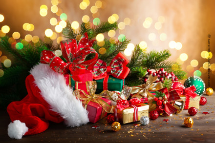 Ein festliches Weihnachtsbild zeigt eine Sammlung von Geschenken, die mit bunten Schleifen verziert sind. Links ist eine rote Weihnachtsmütze mit weißem Pelzbesatz zu sehen, und im Hintergrund befinden sich grüne Tannenzweige.