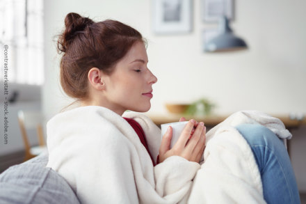 Eine Frau mit braunen Haaren sitzt gemütlich auf einem Sofa, in eine weiße Decke gehüllt, und hält eine Tasse in beiden Händen. Sie hat die Augen geschlossen und wirkt entspannt. Im Hintergrund ist unscharf eine Zimmerdekoration zu erkennen.