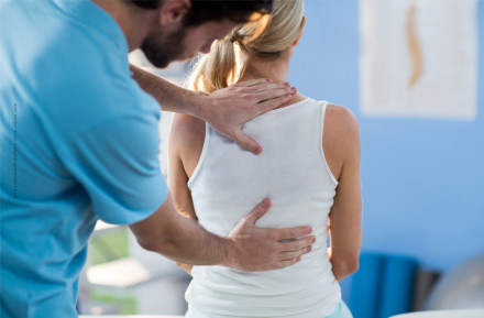 Ein Physiotherapeut behandelt eine Frau, indem er ihr mit seinen Händen den oberen und unteren Rücken massiert. Die Frau sitzt mit dem Rücken zur Kamera und trägt ein weißes Oberteil. Der Therapeut trägt ein hellblaues Hemd.