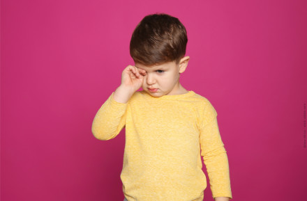 Ein kleines Kind in einem gelben langärmligen Oberteil steht vor einem pinkfarbenen Hintergrund und reibt sich das Auge. Es sieht traurig oder müde aus.