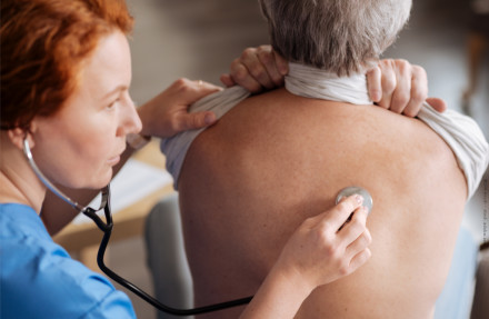 Eine Ärztin hört mit einem Stethoskop den Rücken eines älteren Patienten ab. Der Patient sitzt mit freiem Oberkörper und die Ärztin ist konzentriert bei der Arbeit.