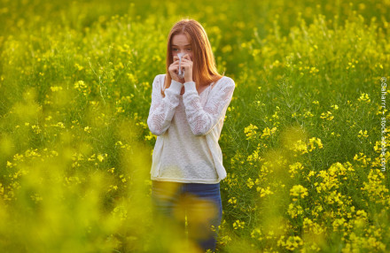 Eine junge Frau mit roten Haaren steht in einem Feld voller gelber Blumen. Sie trägt einen weißen Pullover und blaue Jeans und hält ein Taschentuch vor ihr Gesicht, während sie scheinbar niest oder ihre Nase putzt.