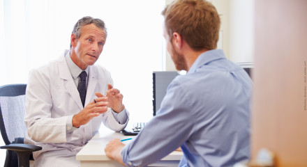 Ein Arzt in einem weißen Kittel spricht mit einem Patienten, der ihm aufmerksam zuhört. Beide sitzen an einem Schreibtisch in einem Büro. Der Arzt gestikuliert und erklärt etwas, während der Patient konzentriert wirkt.