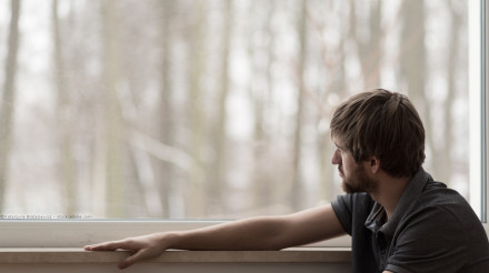 Ein Mann mit kurzen braunen Haaren und Bart sitzt am Fenster und schaut nach draußen. Seine linke Hand ruht auf der Fensterbank. Draußen sind Bäume und ein unscharfer Hintergrund zu sehen. Die Stimmungist  kalt, möglicherweise winterliche Stimmung.