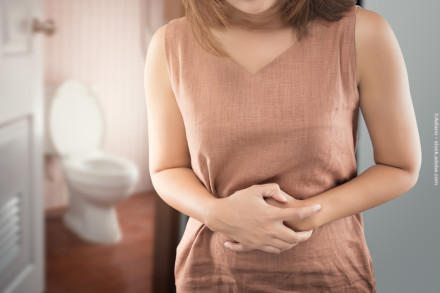 Eine Frau steht vor einem Badezimmer mit einem leicht geöffneten Tür. Im Hintergrund ist eine Toilette sichtbar. Die Frau trägt ein ärmelloses, braunes Kleid und hält sich den Bauch, was auf Bauchschmerzen hinweisen könnte.