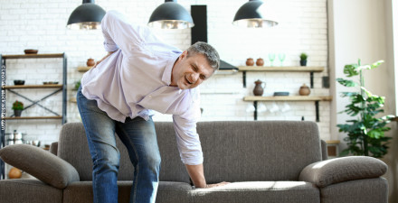 Ein Mann mittleren Alters mit grauen Haaren und einem hellen Hemd steht vor einem Sofa in einem modernen Wohnzimmer. Er hält sich mit schmerzerfülltem Gesichtsausdruck den unteren Rücken und stützt sich mit einer Hand auf das Sofa.