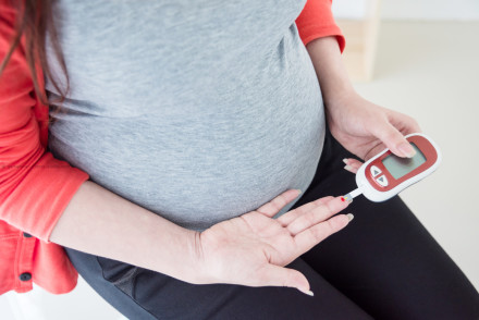 Eine schwangere Frau in grauem T-Shirt und rotem Pullover misst ihren Blutzucker mit einem Glukometer, indem sie einen Bluttropfen von ihrem Finger nimmt.