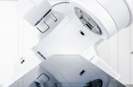 Stellen Sie sich einen modernen medizinischen Linearbeschleuniger in einem strahlentherapeutischen Behandlungsraum vor. Das Gerät ist komplett in Weiß gehalten und wirkt hochtechnologisch.