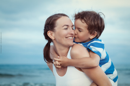 Eine Mutter mit ihrem jungen Sohn auf dem Rücken am Strand. Beide lachen und genießen einen glücklichen Moment zusammen vor dem Hintergrund des Meeres und des bewölkten Himmels.