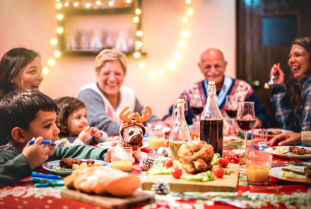 Eine Gruppe von Menschen sitzt an einem festlich gedeckten Tisch und lächelt, während sie eine Mahlzeit genießen. Der Tisch ist mit verschiedenen Speisen, Getränken und Weihnachtsdekorationen geschmückt.