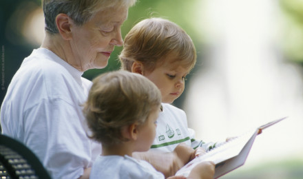 Eine ältere Frau sitzt draußen auf einer Bank und liest zwei kleinen Kindern ein Buch vor. Alle drei tragen weiße T-Shirts und schauen konzentriert auf das Buch. Die Umgebung ist leicht verschwommen, was auf einen Garten oder Park schließen lässt.