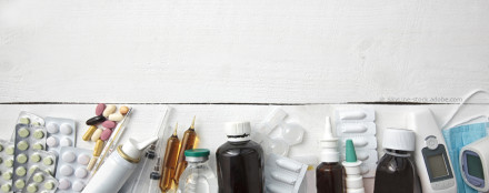 Medikamente und medizinische Geräte auf einem weißen Holzuntergrund. Es gibt verschiedene Tabletten, Sirupflaschen, Nasensprays, Ampullen, eine Spritze, ein Fieberthermometer, ein digitales Blutdruckmessgerät und eine Gesichtsmaske.