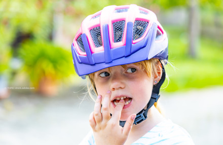 Ein kleines Kind trägt einen lila-rosa Fahrradhelm und steckt sich einen Finger in den Mund. Der Hintergrund ist verschwommen und zeigt grüne Pflanzen.