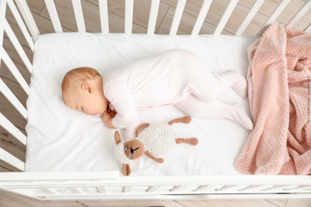 Ein Baby in weißem Strampler schläft auf der Seite in einem weißen Kinderbett. Neben dem Baby liegt ein gestricktes Stofftier in Form eines Schafes. Eine rosa Decke befindet sich am Rand des Kinderbettes.