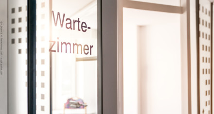 Ein Glasfenster mit der Aufschrift "Wartezimmer". Im Hintergrund ist ein heller Raum zu sehen.