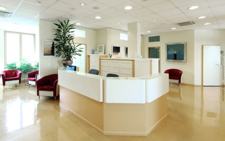 Heller, moderner Empfangsbereich in einem Büro oder einer Klinik. Es gibt eine zentrale Empfangstheke mit zwei Stühlen dahinter, Topfpflanze, Schränke im Hintergrund und mehrere rote Stühle im Wartebereich. Tageslicht kommt durch die Fenster herein.