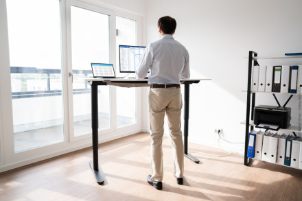 Ein Mann steht vor einem höhenverstellbaren Schreibtisch in einem modernen Büro. Auf dem Schreibtisch befinden sich ein Laptop und ein zusätzlicher Bildschirm mit Tabellen und Diagrammen.