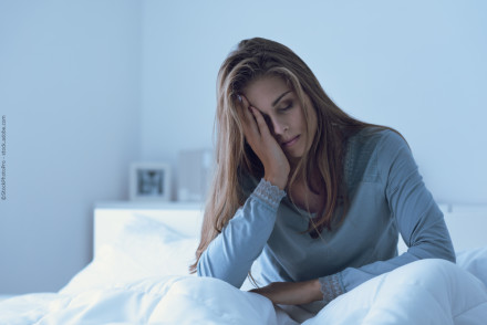 Eine Frau sitzt auf einem Bett und stützt ihren Kopf mit einer Hand. Sie sieht müde und nachdenklich aus. Die Umgebung ist in blauen Tönen gehalten und deutet auf eine kühle Atmosphäre im Raum hin.