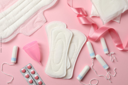 Das Bild zeigt verschiedene Menstruationsprodukte auf einem rosa Hintergrund. Zu sehen sind Damenbinden, Tampons, eine Menstruationstasse, Schmerztabletten und eine rosafarbene Schleife.