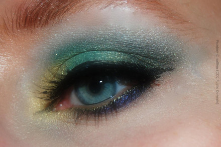 Nahaufnahme eines geschminkten Auges mit blau-grünem Lidschatten, schwarzen Eyeliner und Mascara. Teile des Unterlids sind dunkelblau und das innere Augenlid ist mit einem goldenen Lidschatten versehen.