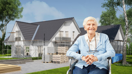 Eine ältere Frau in einem blauen Pullover sitzt im Rollstuhl und lächelt. Im Hintergrund befindet sich ein im Bau befindliches modernes zweistöckiges Haus mit Gerüsten, Baumaterialien und Bäumen. Der Himmel ist klar und blau.