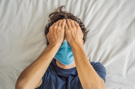 Eine Person liegt auf einem Bett mit einem weißen Laken und trägt eine blaue Gesichtsmaske. Die Person hält sich mit beiden Händen den Kopf und hat die Augen geschlossen, was auf Stress oder Müdigkeit hinweisen könnte.