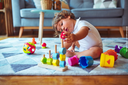 Kleinkind spielt auf einem Teppich in einem Wohnzimmer mit verschiedenen bunten Spielzeugen. Es trägt ein weißes Hemd und ordnet farbige Holzblöcke auf einem Spielzeughalter an.