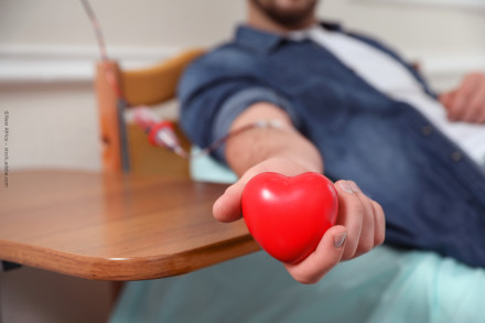 Ein Mann spendet Blut und liegt auf einem Stuhl. Er hält ein rotes Herz aus Gummi in seiner Hand. Ein Schlauch führt von seinem Arm zur Blutsammelvorrichtung.