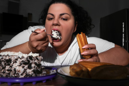 Eine Frau in einem weißen T-Shirt isst Kuchen mit einer Gabel, während sie in der anderen Hand einen Churro hält. Vor ihr auf dem Tisch befindet sich ein großer Stück Kuchen auf einem blauen Teller. Die Szene spielt sich in einer dunklen Küche ab.