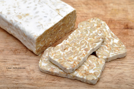 Ein Block Tempeh liegt auf einem Holzschneidebrett. Neben dem Block sind einige Scheiben Tempeh geschnitten und liegen ebenfalls auf dem Brett.