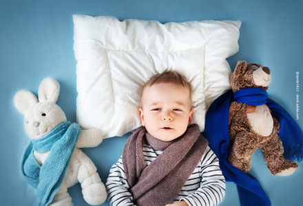 Ein Baby liegt auf einer weißen Kissen auf einem blauen Hintergrund. Es trägt einen gestreiften Pullover und einen grauen Schal. Links vom Baby befindet sich ein weißes Stofftier in Form eines Kaninchens mit einem blauen Schal.