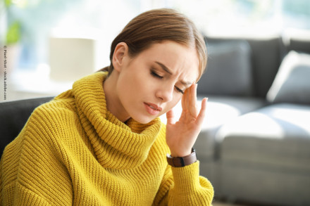 Eine Frau in einem gelben Strickpullover sitzt auf einem Sofa und hält sich den Kopf vor Schmerzen. Sie wirkt, als habe sie Kopfschmerzen. Im Hintergrund ist ein unscharfer Raum zu sehen.
