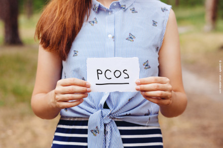 Eine Person hält ein handgeschriebenes Schild mit der Aufschrift "PCOS". Sie trägt ein ärmelloses, blau-weiß gestreiftes Hemd mit Schmetterlingsmustern und einen blau-weiß gestreiften Rock. Im Hintergrund ist eine verschwommene, grüne Outdoor-Umgebung.