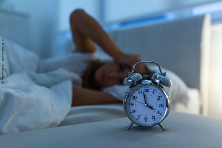 Ein analoger Wecker zeigt 3:55 Uhr an. Im Hintergrund liegt eine Person im Bett und hält sich den Kopf. Die Szene ist dunkel, es scheint früher Morgen oder späte Nacht zu sein.