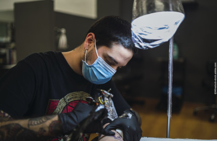 Ein Tätowierer mit schwarzem T-Shirt und Einweghandschuhen tätowiert den Arm einer Person. Er trägt eine blaue Gesichtsmaske und konzentriert sich auf seine Arbeit, während eine Lampe über ihm leuchtet.