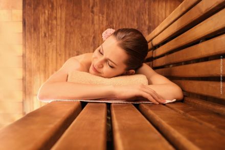 Eine Frau liegt entspannt auf einem Handtuch in einer Sauna und hat die Augen geschlossen. Sie hat eine Blume im Haar und wirkt ruhig und zufrieden. Im Hintergrund sind Holzwände und Sitzbänke zu sehen.