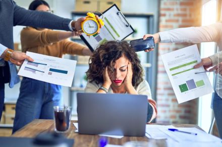 Eine gestresste Frau sitzt vor einem Laptop und hält sich den Kopf. Um sie herum halten mehrere Personen Dokumente, ein Telefon und eine Uhr in ihre Richtung. Die Szene vermittelt einen Eindruck von Überforderung und Arbeitsstress.