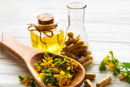 Ein Holzlöffel mit gelben Blüten und grünen Blättern liegt auf einer weißen Holzoberfläche. Im Hintergrund steht eine kleine Glasflasche mit Korkverschluss, gefüllt mit einer gelben Flüssigkeit, sowie ein weiteres Glas mit Kapseln.