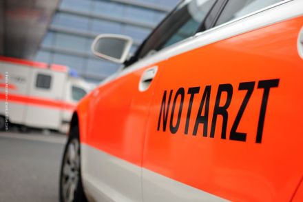 Ein orange-weißes Notarztfahrzeug mit der Aufschrift "NOTARZT" auf der Seite ist in Nahaufnahme zu sehen. Im Hintergrund befindet sich ein weiterer Krankenwagen vor einem modernen Gebäude.