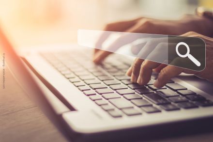 Eine Person tippt auf einer Laptop-Tastatur, während sie eine virtuelle Suchleiste mit einer Lupe verwendet. Das Bild ist in warmen, gedämpften Farben gehalten.