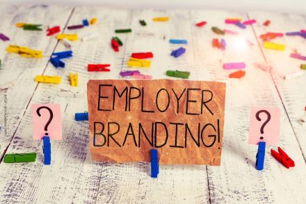 Ein hölzerner Hintergrund, auf dem viele bunte Wäscheklammern verstreut liegen. In der Mitte des Bildes befindet sich ein beschriftetes Schild mit der Aufschrift "EMPLOYER BRANDING!" in großen Buchstaben, gehalten von blauen Wäscheklammern.