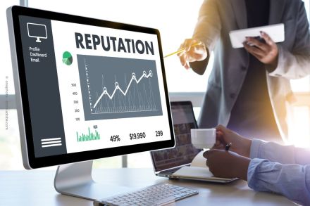 Ein Computerbildschirm zeigt ein Diagramm mit dem Titel "REPUTATION". Es gibt eine Grafik mit ansteigender Kurve und Kennzahlen darunter. Eine Person mit einem Stift deutet auf den Bildschirm, während eine andere eine Tasse und ein Notebook hält.