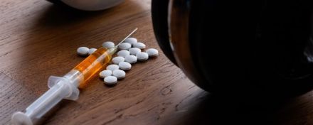 Nahaufnahme einer Spritze mit orangener Flüssigkeit und weißen Tabletten auf einem Holztisch. Im Hintergrund sind verschwommen Hanteln zu sehen, was auf den möglichen Missbrauch von Steroiden hinweist