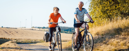 Ein älteres Paar fährt bei sonnigem Wetter auf einem ländlichen Weg Fahrrad. Die Frau trägt ein orangefarbenes Shirt, der Mann ein hellblaues. Im Hintergrund Felder und Bäume, die eine entspannte und gesunde Outdoor-Aktivität darstellen.